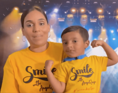 Picture of Angel Jayce and his mother. Both are wearing yellow shirts that say the word smile on it. The mom is wearing earrings, while Angel Jayce has a blue collar around is neck. Both have brown hair, and they are in front of a stage with many bright lights