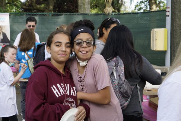 Picture of two young ladies smiling. One has black hair and one has brown hair. One is wearing a light pink shirt with a Nike headband and glasses and the other has a hooded sweat shirt that says Harvard on it. Both are outside surrounded by a group of people