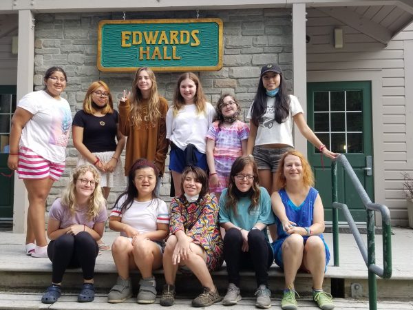 myFace - Camp Reflections 2021 Picture of young teens smiling, outside in a camp setting. One row of kids is sitting down on steps while the second row is standing behind them. They are in front of a gray building that has a green and yellow sign that says Edwards Hall