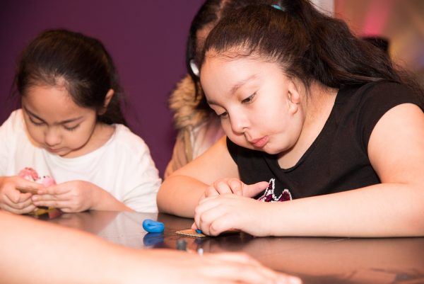 myFace - Kids At Holiday Party 2019 Picture of two young girls with long dark brown hair, one is wearing a black shirt while the other is wearing a white shirt. Both are playing with blue play dough and cracker