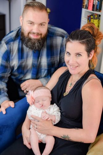 myFace - Mauriellos_Ashley, Tommy, Harlyn Picture of parents with a baby, Mother has long brown and blond hair, wearing a black dress with two green necklaces, she is holding a baby that is wearing a white onesie and a pink flower headband. The father has short brown hair, a brown and gray beard, and is wearing a blue plaid shirt