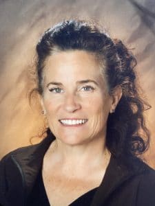 A picture of Amy Schefer with a grayish-orange gradient background. Amy is smiling and has long, dark wavy hair. She is Caucasian and is wearing a dark, collared v-neck shirt.