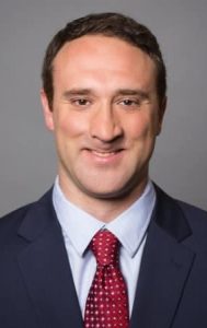 Picture of Max Singer against a plaiine grya background. Max is a Caucasian male and has a facial difference, he has short dark hair and he is smiling. He is wearing a gark suit jacket with a light blue collared shirt and a red tie with small, white polka dots on it