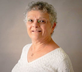 Picture of Luci Capo Rome against a gray-beige background. Luci has short, curly, white-gray hair. She is wearing glasses, and she is smiling. She is also wearing a necklace and a white v-neck blouse.