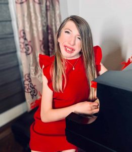 Picture of Grace Novacheck smiling with long dark blonde hair and wearing a bright red dress with ruffles on the soldiers, sitting at a black piano