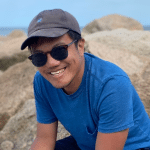 photo of a young man in a hat and sunglasses sitting on a rock outside in the sun and smiling